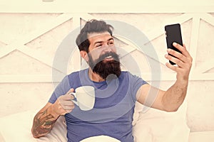 Good morning. Bearded man using mobile technology in bed. Handsome guy talking phone and drinking coffee at home. Happy