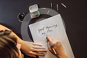 Good morning. Calligrapher Young Woman writes phrase on white paper. Inscribing ornamental decorated letters