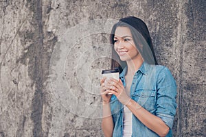 Good morning! Cheerful pretty young serene brunette lady is having hot tea near concrete wall outside, smiling, wearing cozy casua