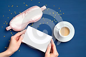 Good morning concept. Cup of coffee, sleep eye mask and female hands holding blank paper card mockup on dark blue background. Flat