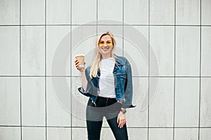 Good morning! Stylish happy young blonde woman wearing stylish jeans jacket, fashion yellow glasses. Cheerful Female holds coffee