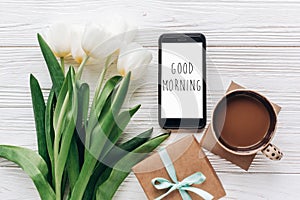 good morning text sign on phone screen and stylish gift and tulips and coffee on white wooden rustic background. flat lay with fl