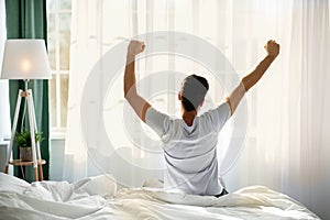Good morning. Young man waking up in bed and stretching his arms, facing the window, back view