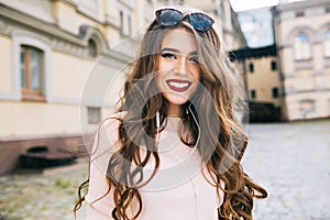 Good-natured girl with long curly hair and vinous lips is smiling to camera in the city