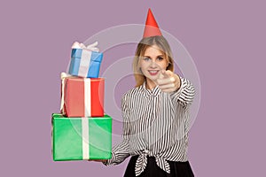 Good-natured happy woman with funny party cone on head pointing to camera and holding many gift boxes photo