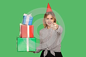 Good-natured happy woman with funny party cone on head pointing to camera and holding many gift boxes photo