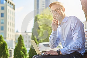 Good news. Young happy businessman talking by phone and and smiling while sitting on the bench outdoors and working