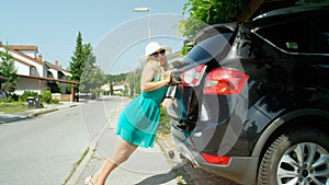 Goofy woman in sundress leans on the car trunk stuffed with baggage to close it.