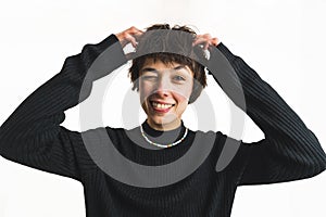 Goofy young girl sticking her tongue out, blinking at camera and touching her short hair. Indoor studio portrait. White