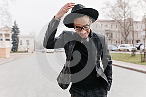 Gorgeous african man posing with shy smile on city background. Stylish black guy in hat standing on the road with