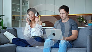 Gorgeous African woman in headphones, dancing singing sitting on couch next to her boyfriend, focused on working on