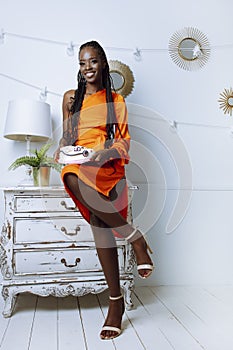 Gorgeous afro american woman in orange dress sit on vintage dresser with holiday cake in white decorated studio. Smiling
