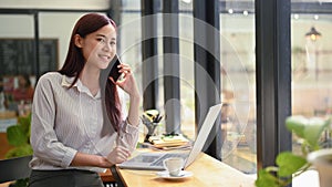 Gorgeous asian business woman having cell phone conversation while sitting front open laptop computer in coffee shop