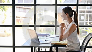 Gorgeous Asian businesswoman sits at her office desk, looking out the window, daydreaming