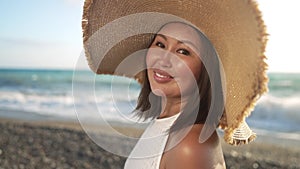 Gorgeous Asian middle aged woman in straw hat posing on Mediterranean sea beach in slow motion. Portrait of beautiful
