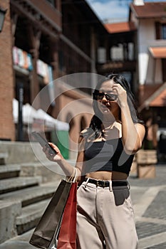 Gorgeous Asian woman enjoys shopping in the city, walking while holding her phone and shopping bags