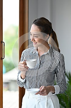 Gorgeous asian woman holding coffee cup and looking out of glass window.