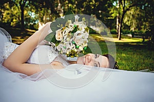 Gorgeous beautiful bride in a white dress lying on the green grass in the Park with a bouquet of flowers in her hands