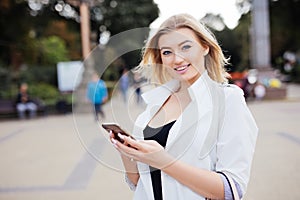 Gorgeous beautiful young woman with blonde hair messaging on the smart-phone at the city street background.
