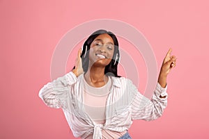 Gorgeous black lady with headset listening to music and dancing with closed eyes, pointing aside on pink background