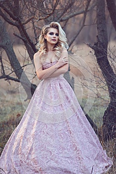 Gorgeous blond lady with luxuriant hairstyle in brocade ball gown standing at the dry leafless tree