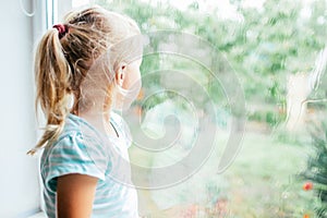 A gorgeous blonde little girl with ponytale staring out of the window on a wet, cold rainy summer day