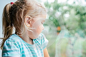 A gorgeous blonde little girl with ponytale staring out of the window on a wet, cold rainy summer day