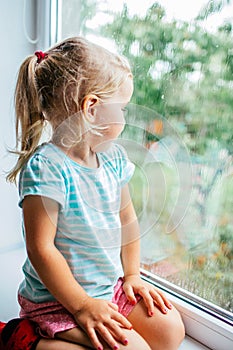 A gorgeous blonde little girl with ponytale staring out of the window on a wet, cold rainy summer day