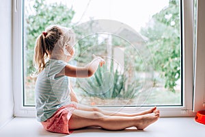 A gorgeous blonde little girl with ponytale staring out of the window on a wet, cold rainy summer day and showing somewhere