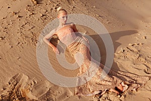 Gorgeous blonde woman in gorgeous golden dress posing in the desert, lying on the sand at sunset. Full length portrait