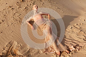 Gorgeous blonde woman in sexy golden dress posing in the desert, lying on the sand.