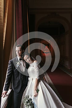 Gorgeous bride in amazing dress with bouquet and stylish groom posing at window in luxury room in hotel. rich wedding couple