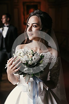 Gorgeous bride in amazing dress with bouquet and stylish groom posing at window in luxury room in hotel. rich wedding couple