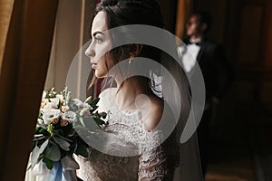 Gorgeous bride in amazing dress with bouquet and stylish groom posing at window in luxury room in hotel. rich wedding couple