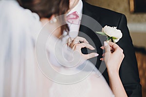 Gorgeous bride in amazing gown putting on white peony boutonniere groom suit. Beautiful woman getting ready in the morning.