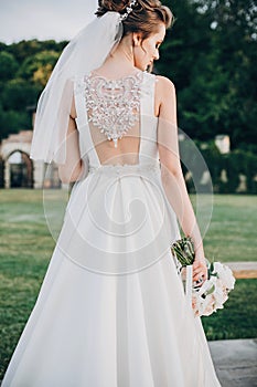 Gorgeous bride in amazing gown walking in evening park. Bride in stylish dress posing with bouquet, back view