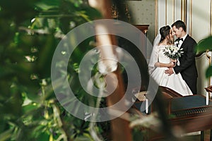 Gorgeous bride with bouquet and stylish groom gently hugging among green leaves in luxury room in hotel. rich wedding couple