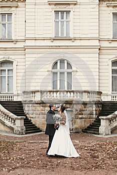 Gorgeous bride with bouquet and stylish groom walking at old castle in european city street in autumn time. happy wedding couple