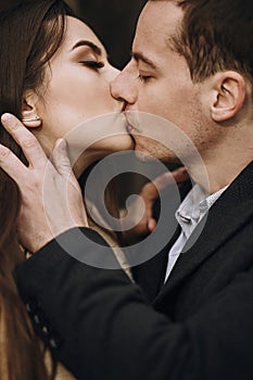 Gorgeous bride in coat and stylish groom kissing under trees in winter forest. happy wedding couple gently embracing in winter