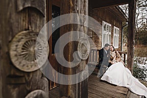 Gorgeous bride in coat and stylish groom sitting at old wooden house in winter forest. happy wedding couple gently hugging on