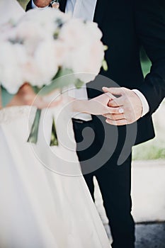 Gorgeous bride and groom gently holding hands in evening park. Stylish wedding couple embracing, holding hands during walk in