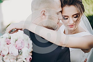 Gorgeous bride and groom gently hugging in sunset light. Beautiful stylish wedding couple embracing in summer park. Sensual