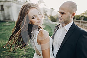 Gorgeous bride and groom hugging and dancing in evening sunlight near old castle in park. Stylish wedding couple gently embracing