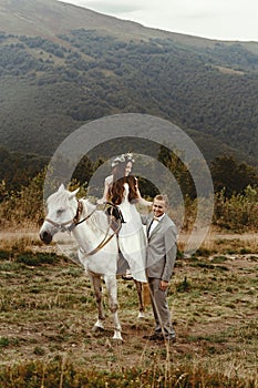 Gorgeous bride riding a white horse and stylish groom, boho wed