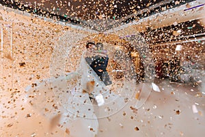 Gorgeous bride and stylish groom dancing under golden confetti at wedding reception.