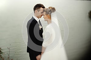 Gorgeous bride and stylish groom gently hugging at sandy beach lake, beautiful moment, luxury wedding