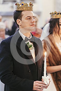 Gorgeous bride and stylish groom in golden crowns, holding candles with light in  church during wedding ceremony.  spiritual