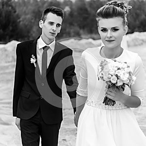 Gorgeous bride and stylish groom holding hands and looking at sandy beach lake, black and white photo, luxury wedding