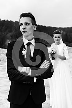 Gorgeous bride and stylish groom holding hands and looking at sandy beach lake, black and white photo, luxury wedding