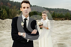 Gorgeous bride and stylish groom holding hands and looking at sandy beach lake, true emotions, luxury wedding